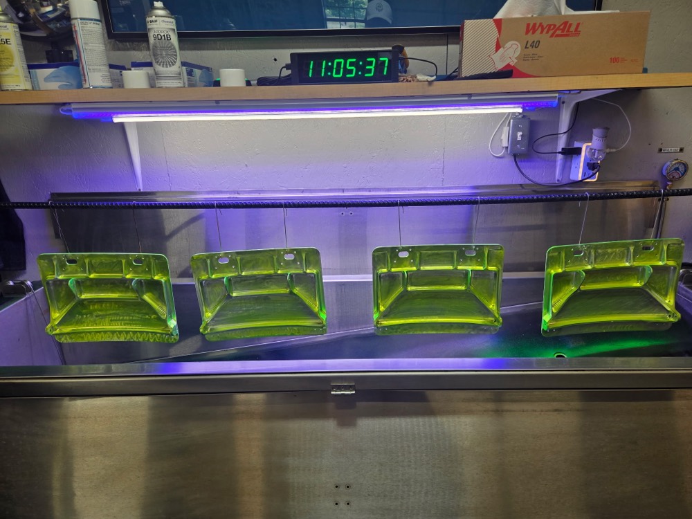 Parts coated in fluorescent penetrant on the inspection line, with the shop clock timing the dwell