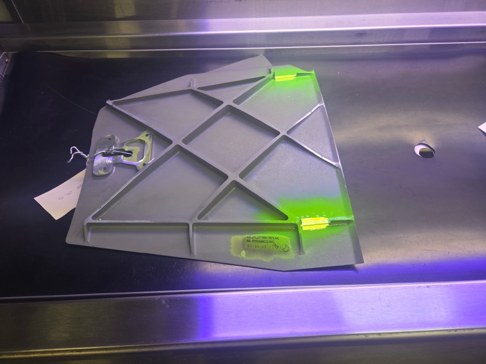 Aircraft panel showing fluorescent penetrant indications under UV-A light, with inspection tag attached